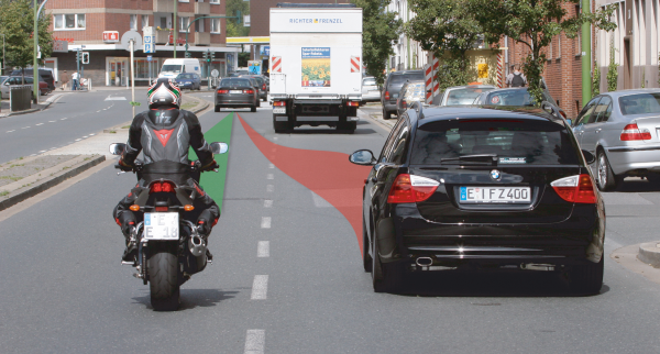 Motorradfahrer und Auto fahren nebeneinander, das Auto setzt zum Spurwechsel an.